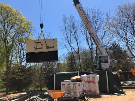 Crane lifting a large wooden crate at a wooded construction site with pallets and tarps nearby