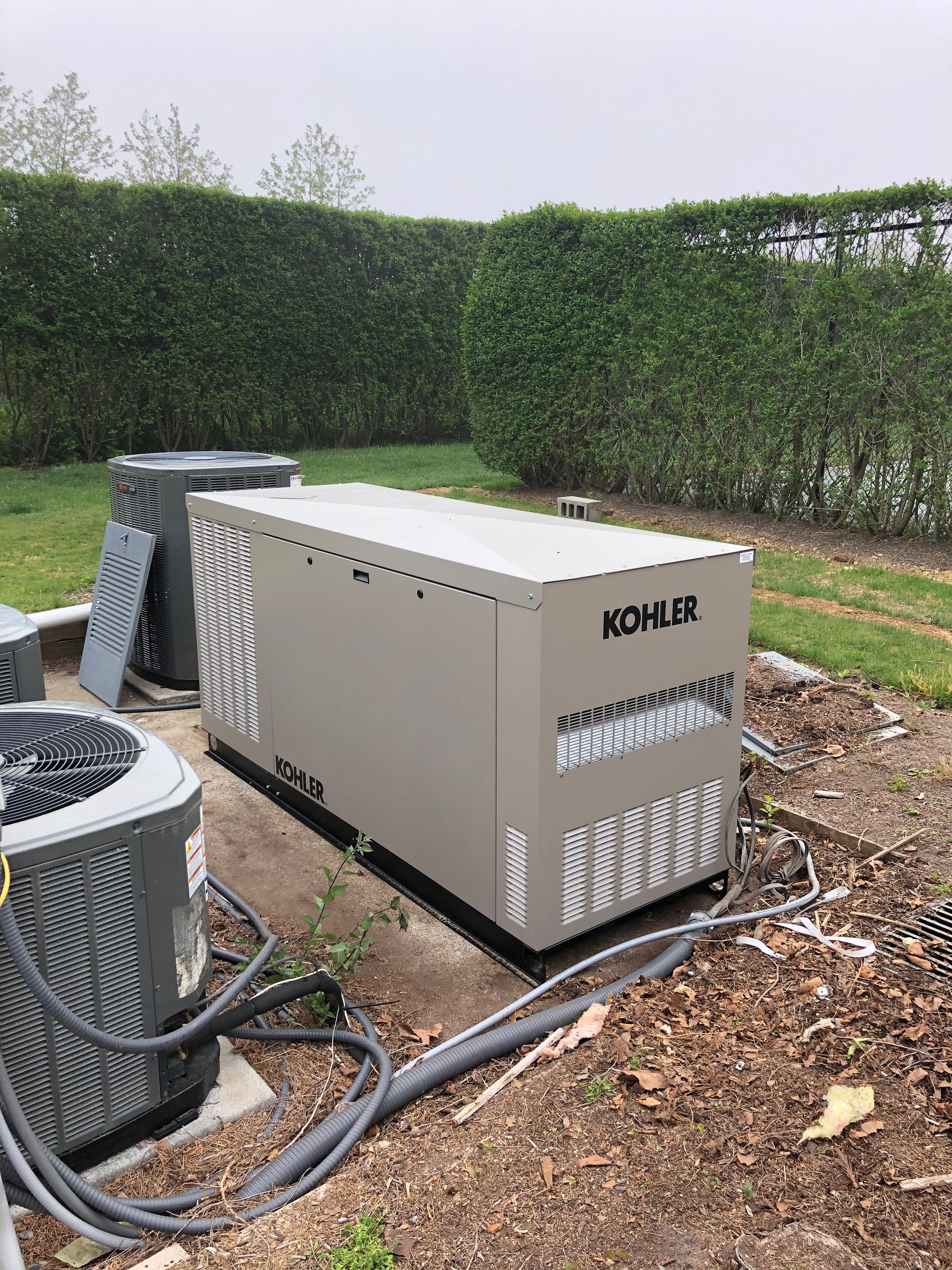 Outdoor Kohler generator on a patio beside an air-conditioning unit, with hoses and a hedge background