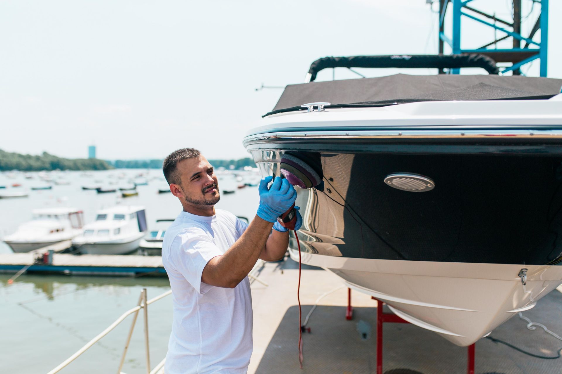 Man polishing a black boat hull with a power buffer at a dock.