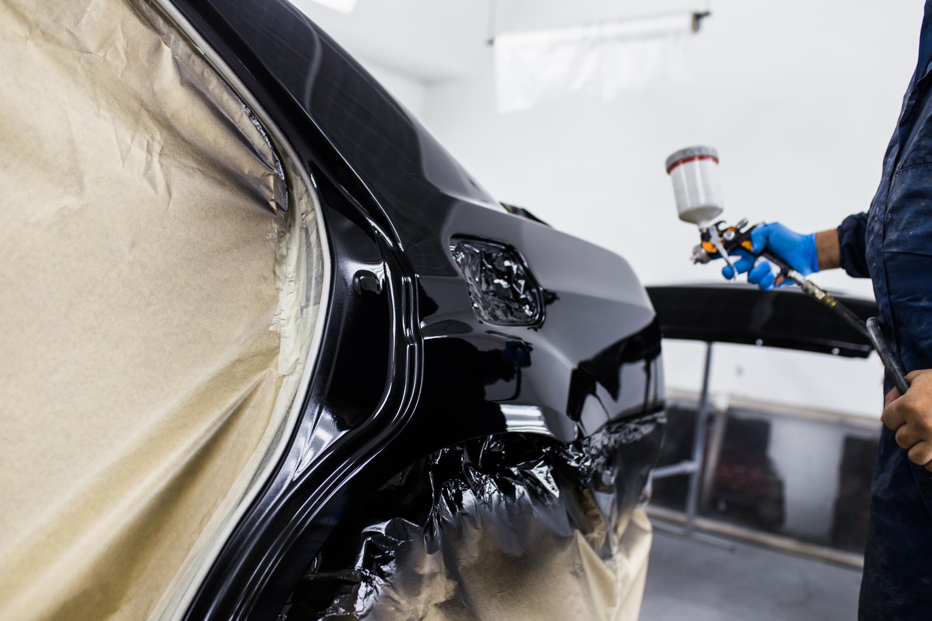 Person in blue gloves spraying black paint on a car's bumper in a shop.