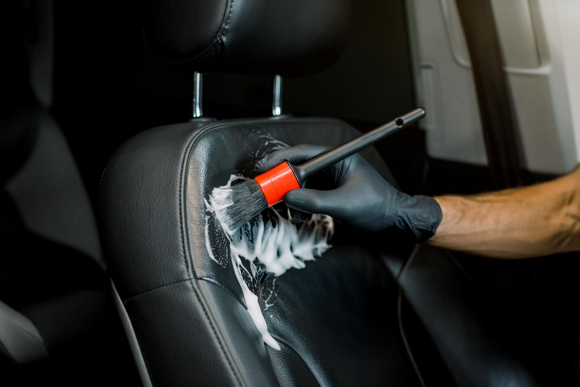 Person in black gloves cleaning a black car seat with a brush and foamy cleaner.