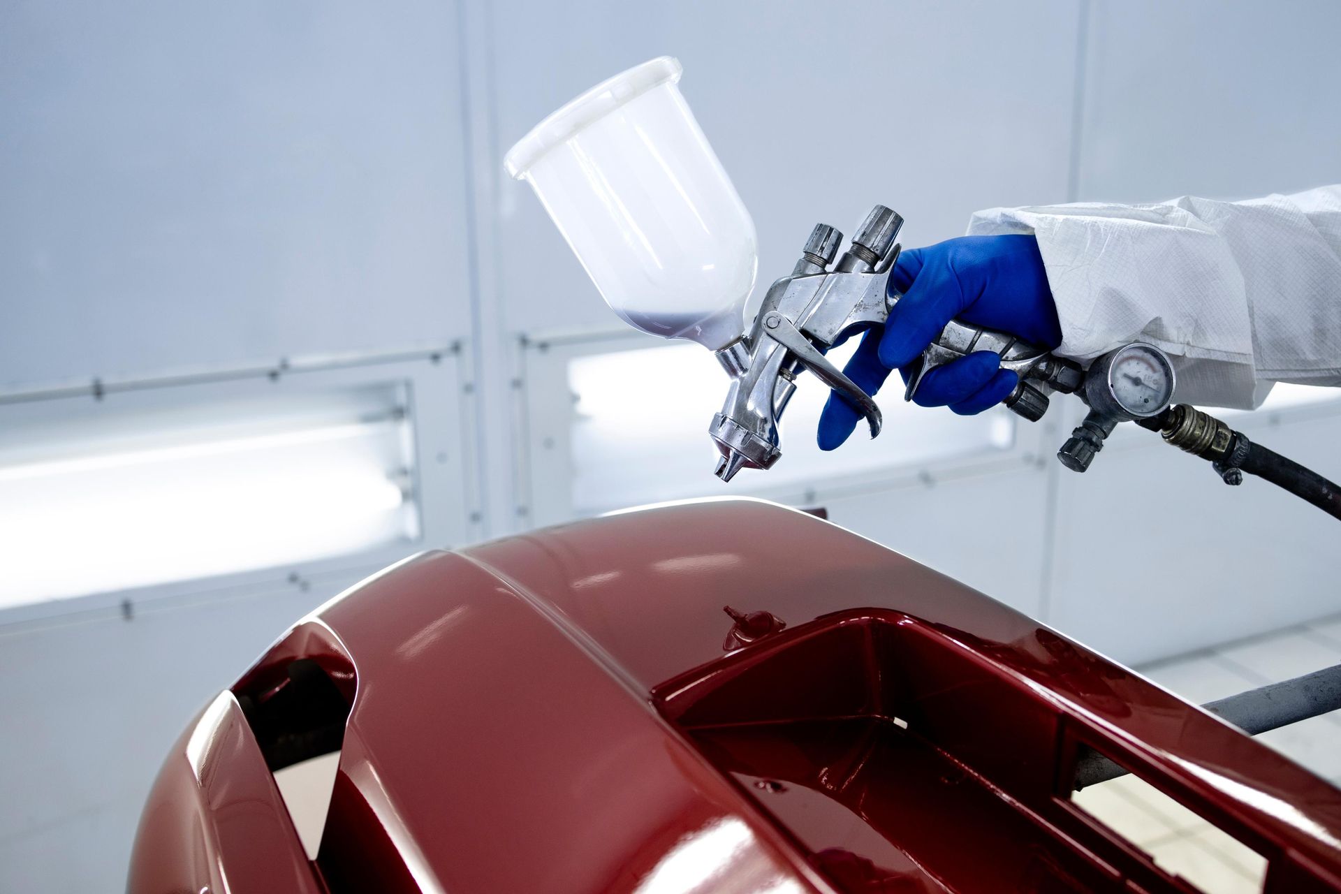Person in blue gloves and white coat sprays red paint onto a car bumper in a paint booth.
