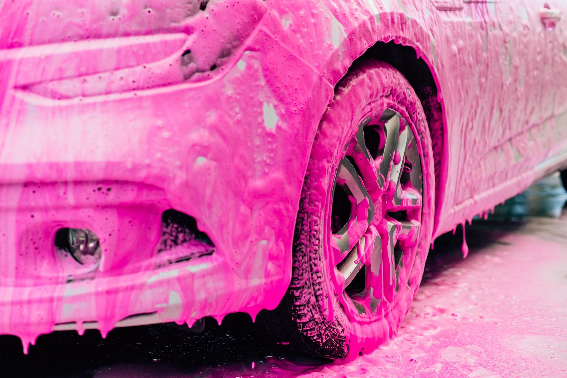 Car covered in vibrant pink foam at a car wash, with a close-up of the front wheel.