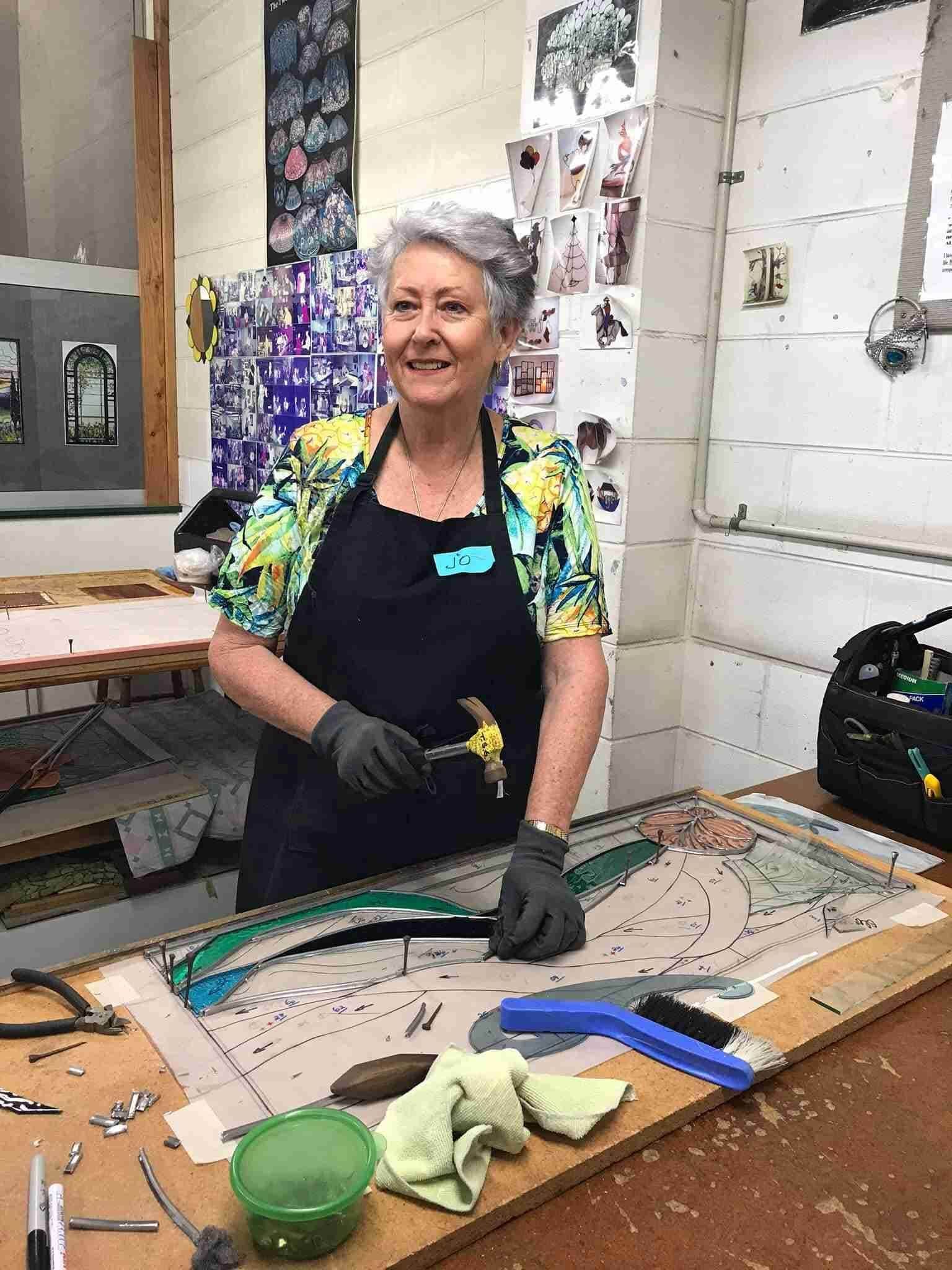 A Woman Is Standing At A Table In A Workshop Holding A Hammer — Townsville Stained Glass In Aitkenvale, QLD