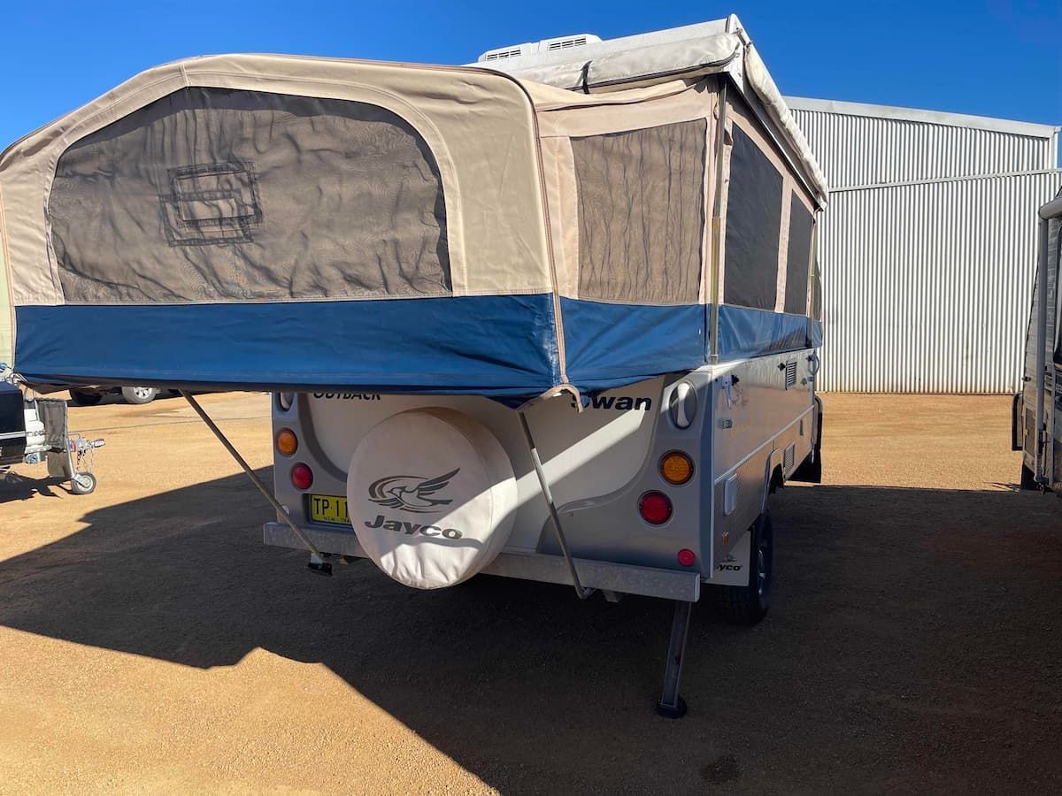 Small White Camper Trailer with Beige Pop Up Tent and Blue Trim Parked on Dirt Near a Metal Building — B & C Caravan Service Pty Ltd In Dubbo, NSW