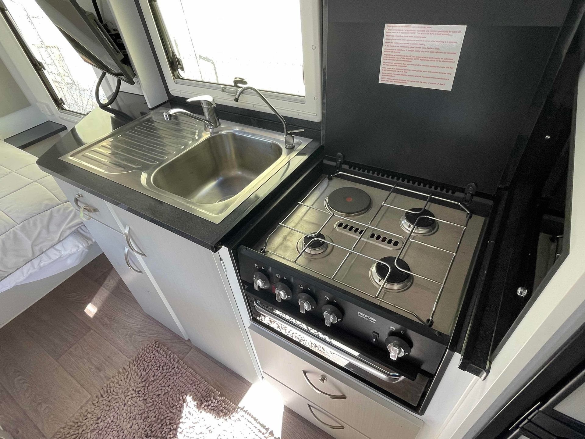 Compact RV Kitchen with Sink, Stove, Oven, and White Cabinets by A Window — B & C Caravan Service Pty Ltd In Dubbo, NSW