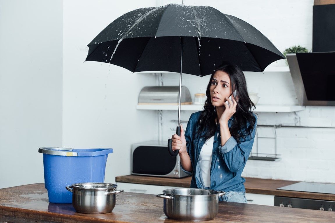 Woman in kitchen uses umbrella to catch water leaking from the ceiling, talking on the phone, with pots and a bucket to collect the water.
