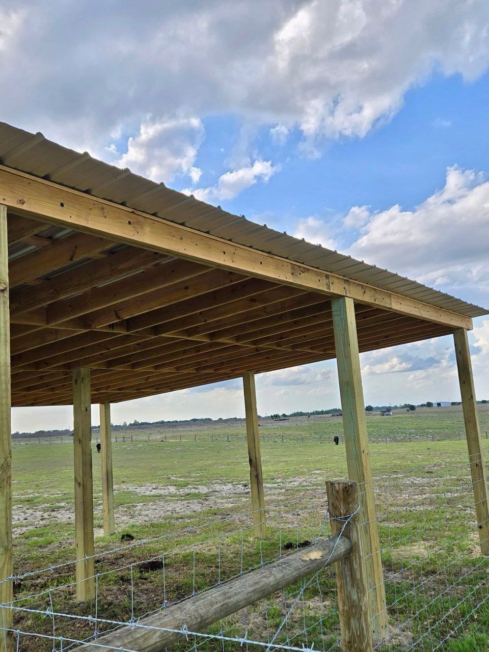 Wooden farm shelter with a corrugated roof in a grassy field under a cloudy blue sky