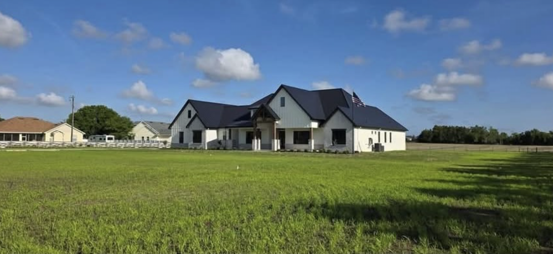 Large white house with a dark roof beside a green field under a blue sky with clouds