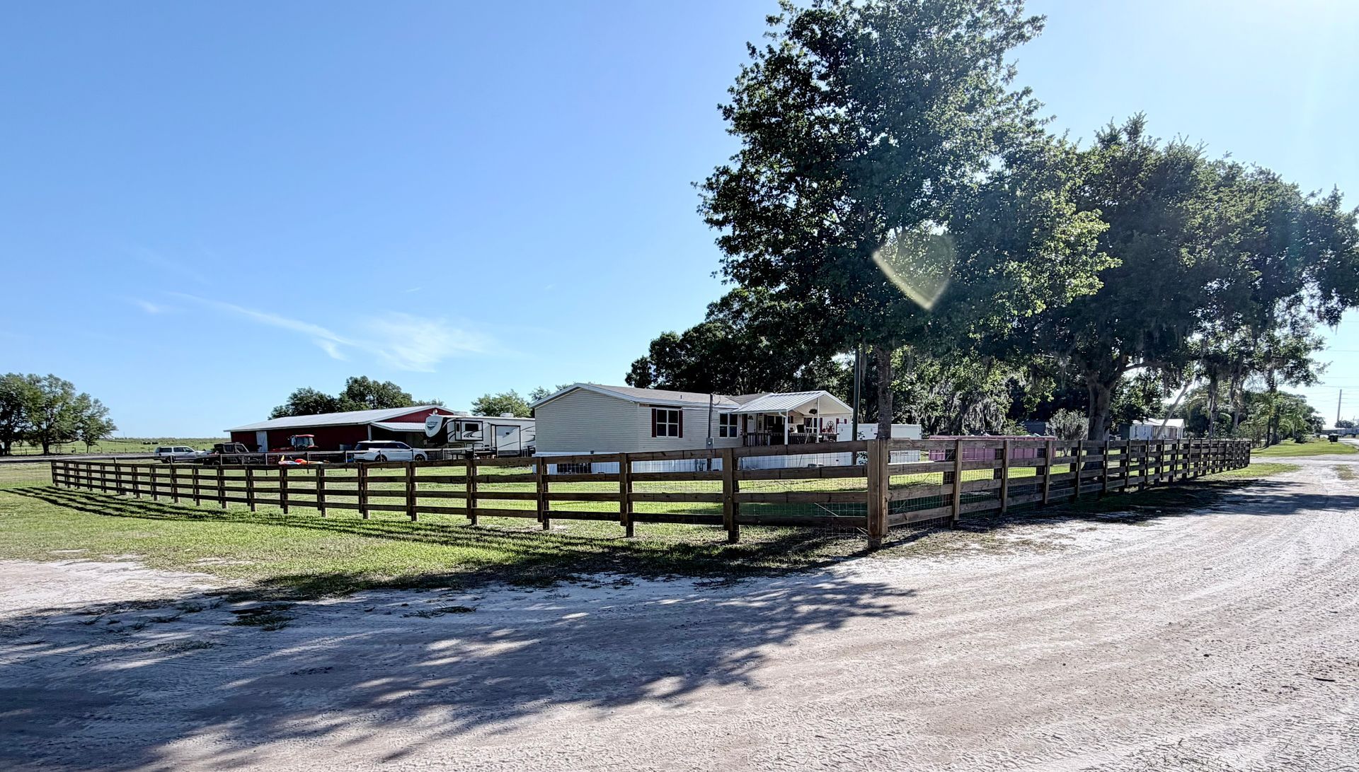Rural property with white house, fenced yard, large tree, and gravel road under a clear blue sky