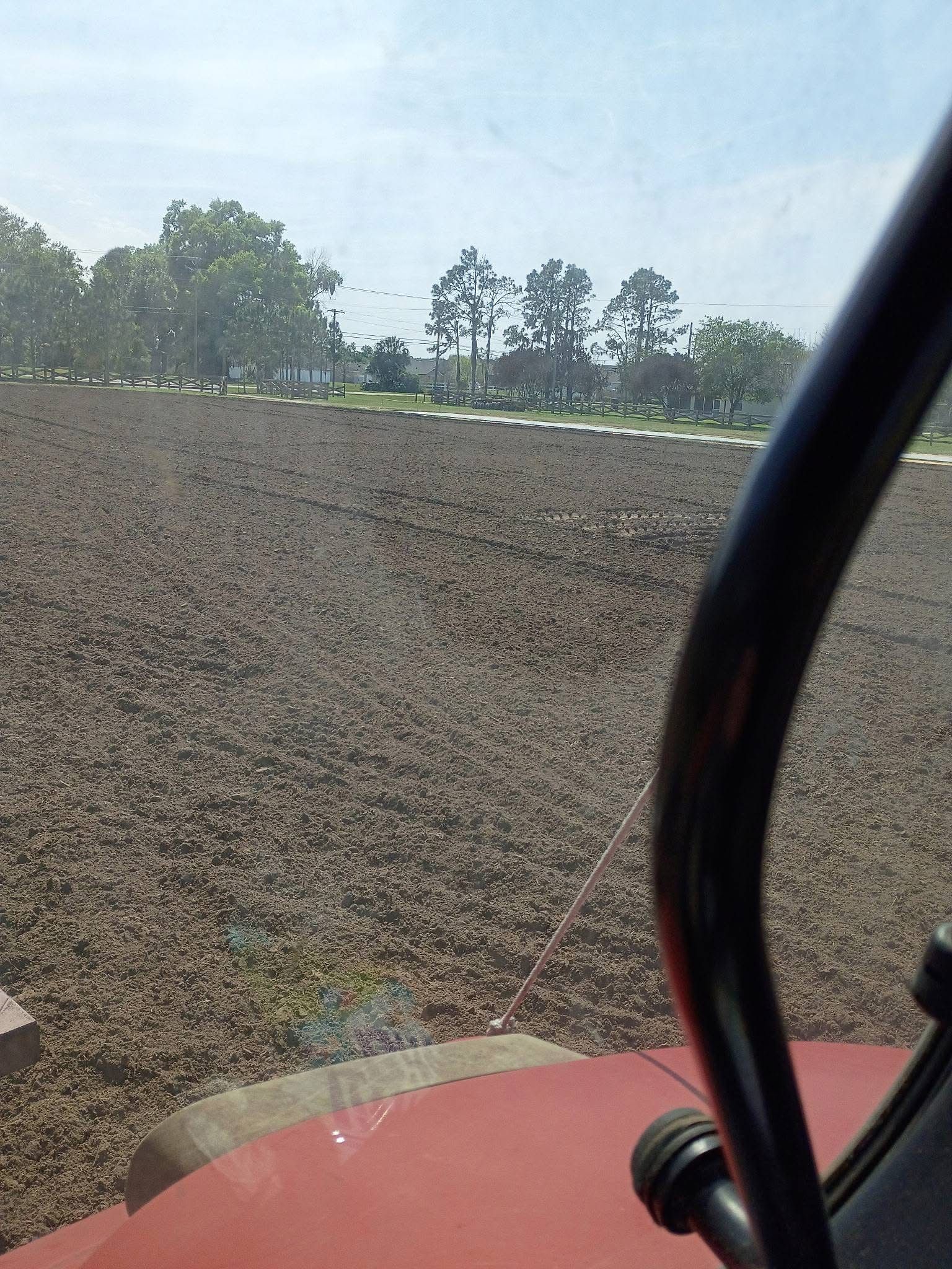 Red vehicle hood and mirror framing a plowed field with trees in the distance under a clear sky