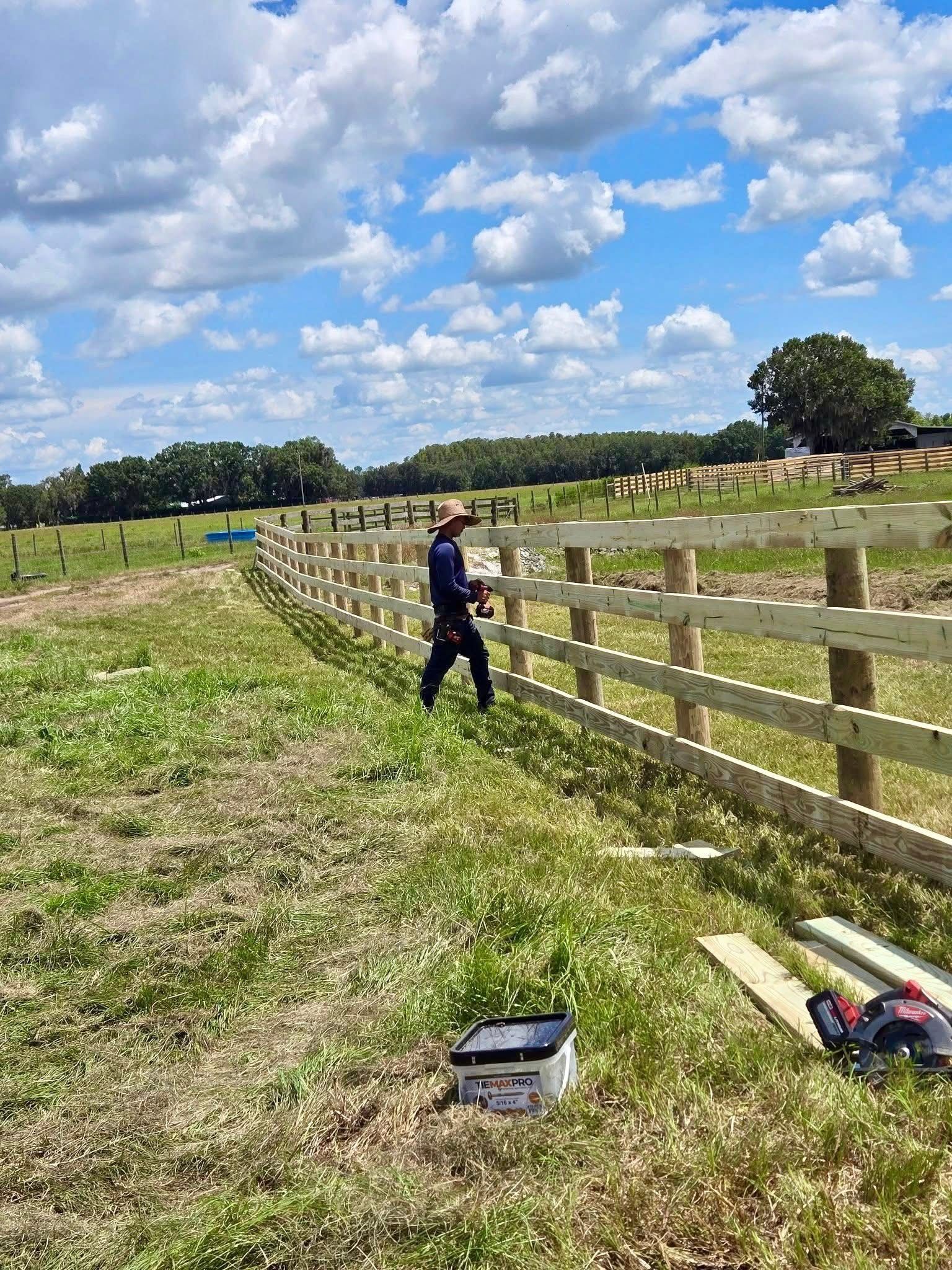 Person working beside a wooden fence in a grassy field under a bright blue sky with clouds.
