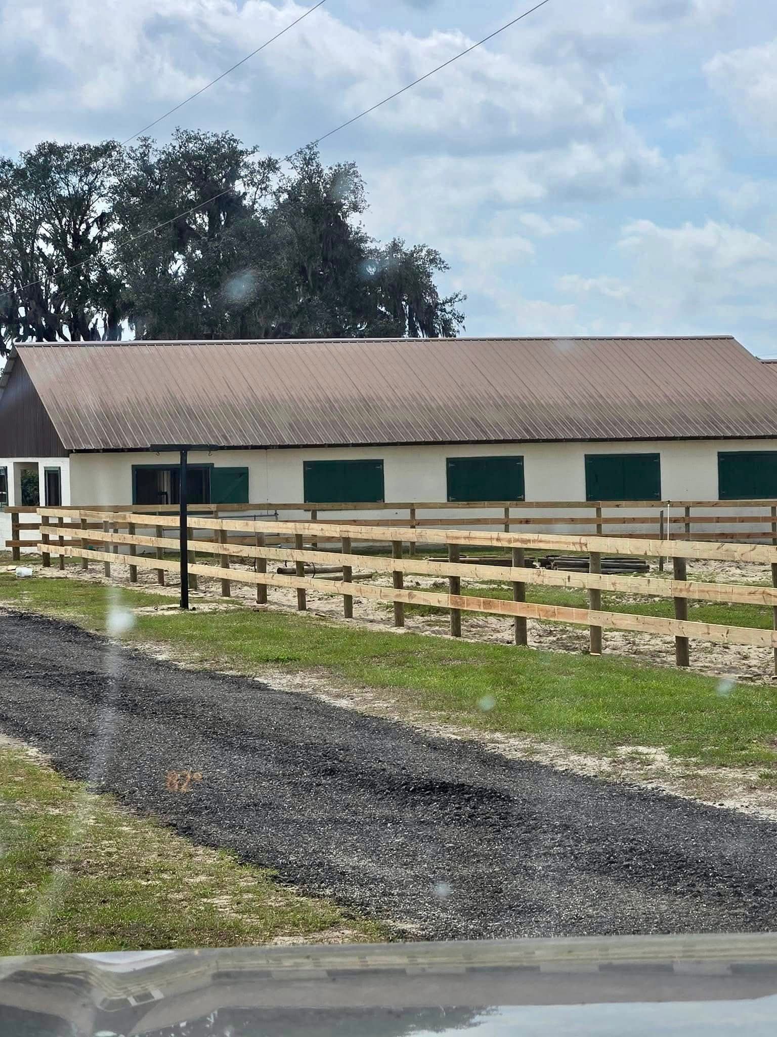 Ranch-style barn with green shutters and white fence beside a gravel drive under cloudy sky