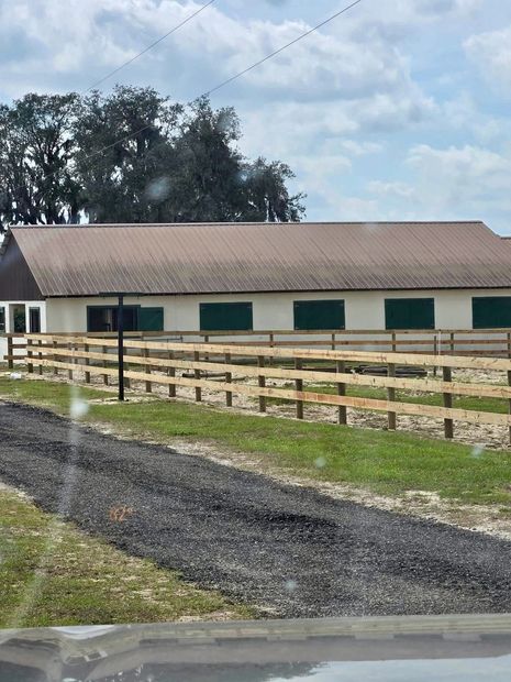 Ranch-style barn with green shutters and white fence beside a gravel drive under cloudy sky