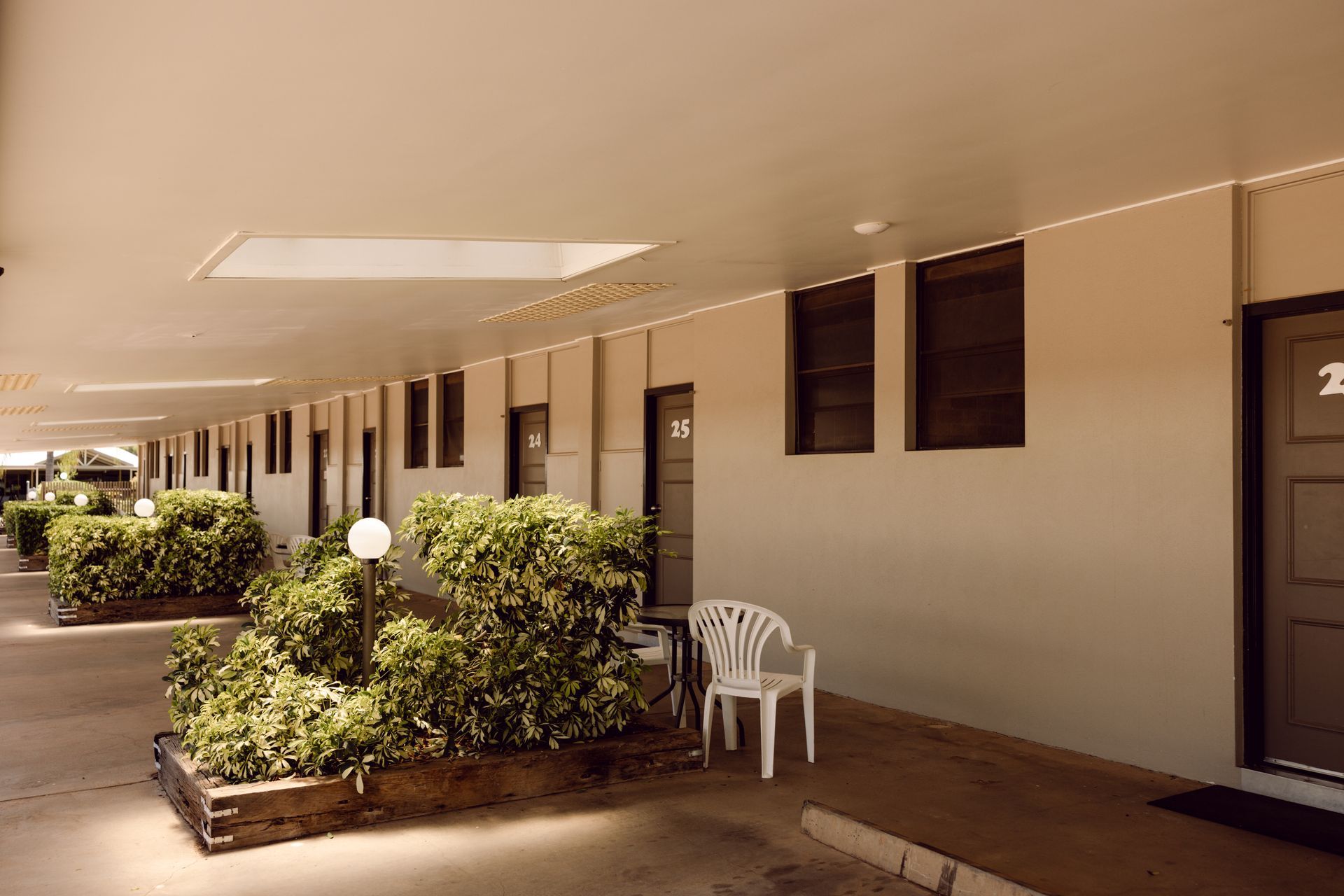 Motel walkway with beige doors, potted shrubs, and white chairs under a covered corridor.