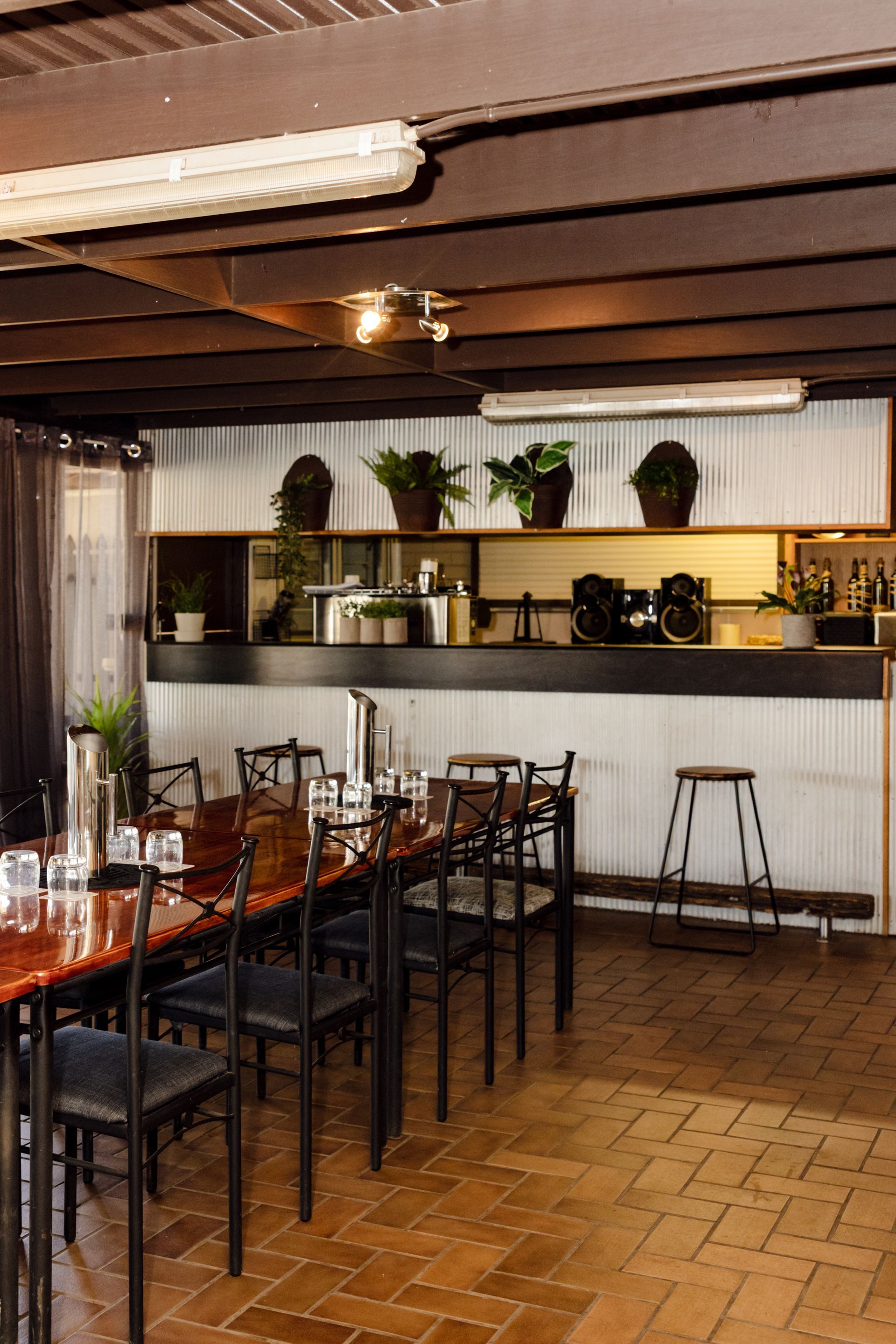 Empty restaurant dining area with wooden tables, black chairs, potted plants, and a brick floor