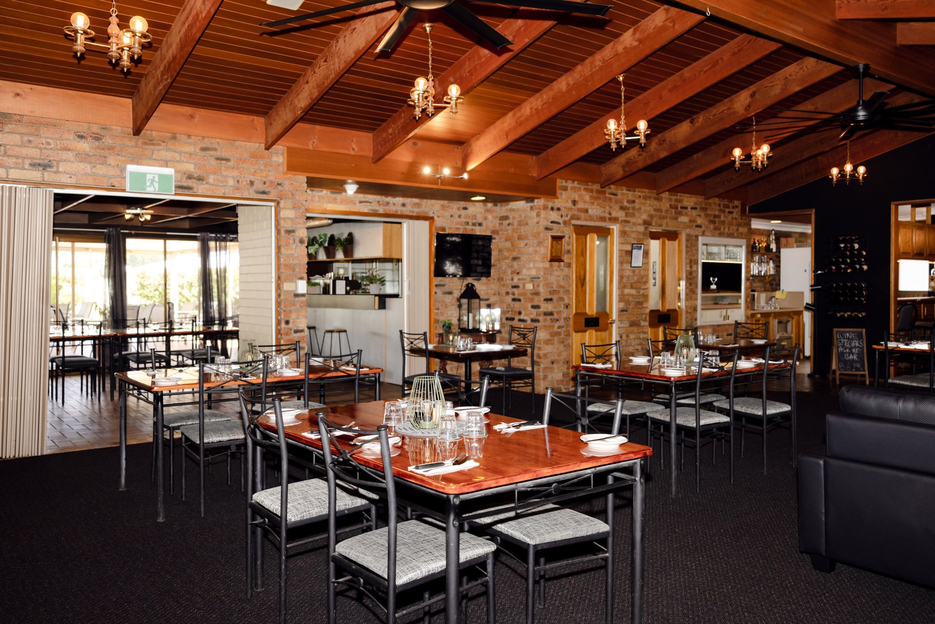 Empty rustic restaurant dining room with wood beams, brick walls, and set tables under warm chandeliers