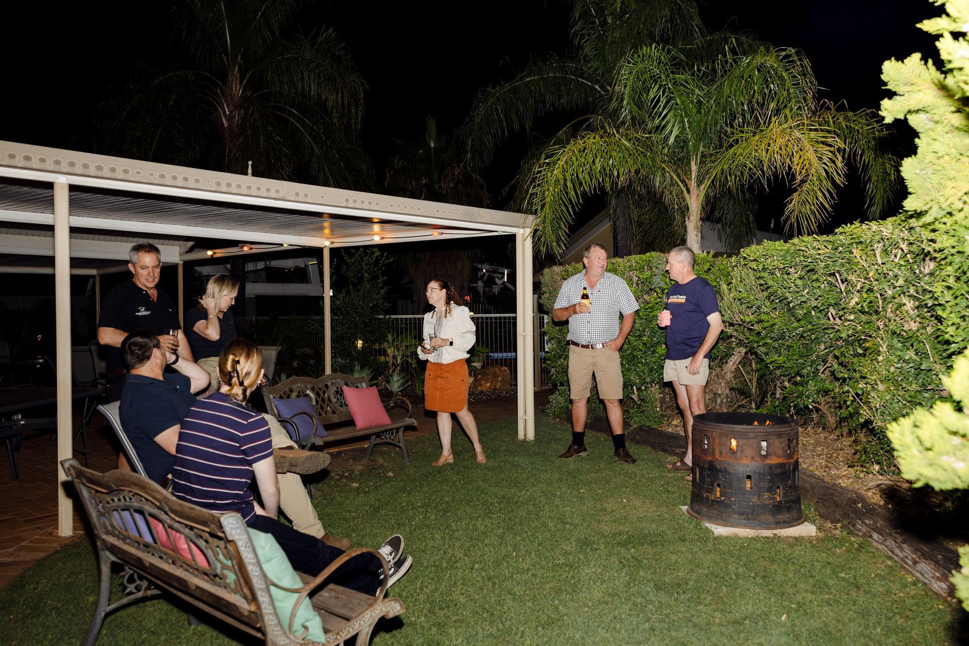 People gathered in a backyard at night, chatting near a covered patio and a fire pit.