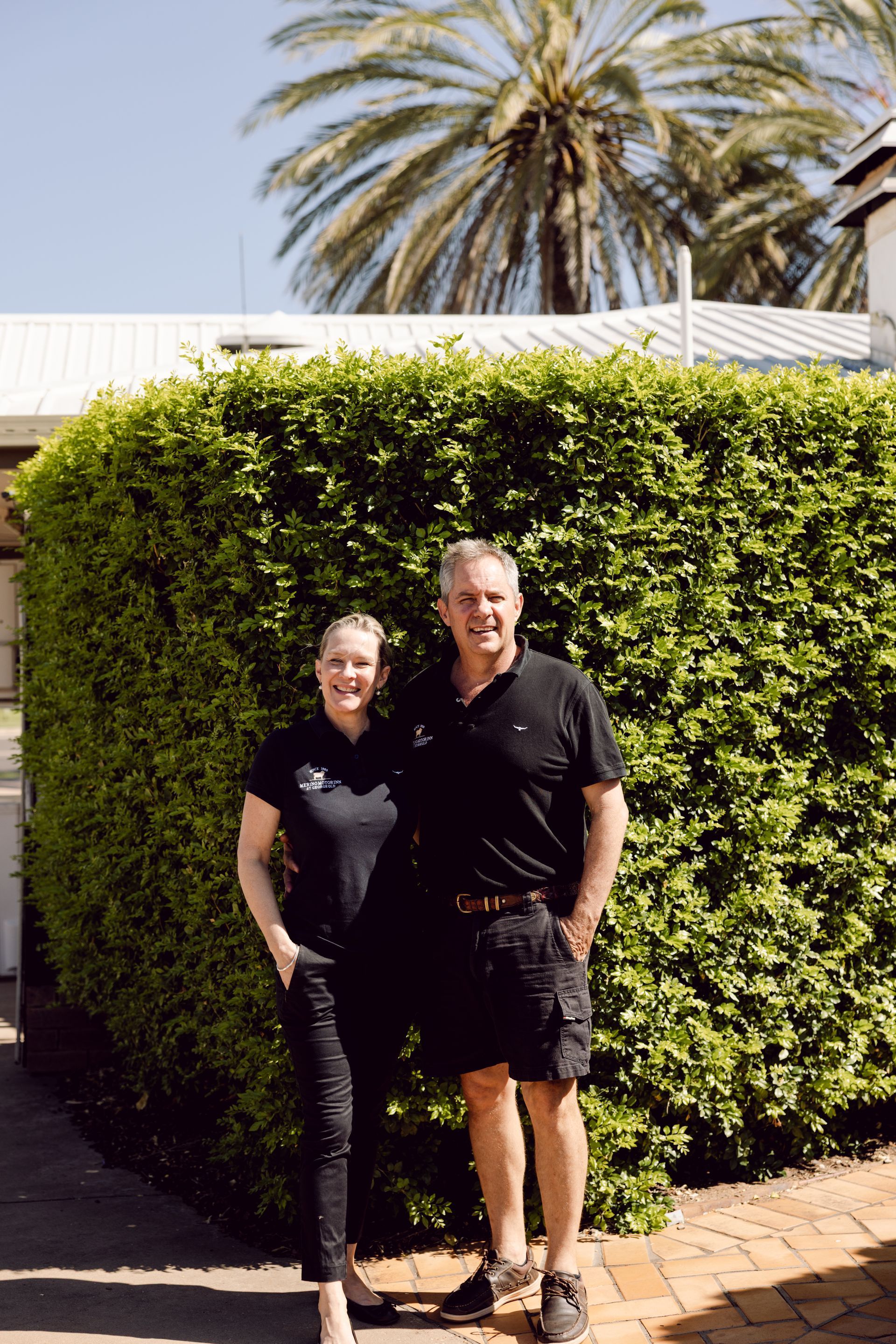 Two people standing together in front of a tall green hedge with a palm tree behind them