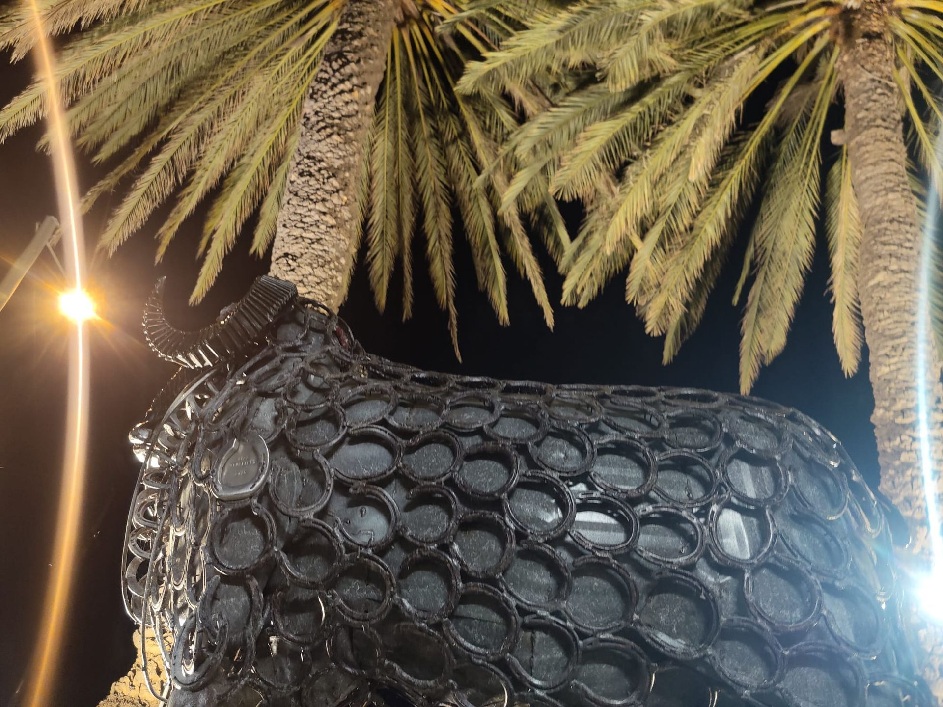 Black textured animal sculpture under palm trees at night, lit by bright streetlights