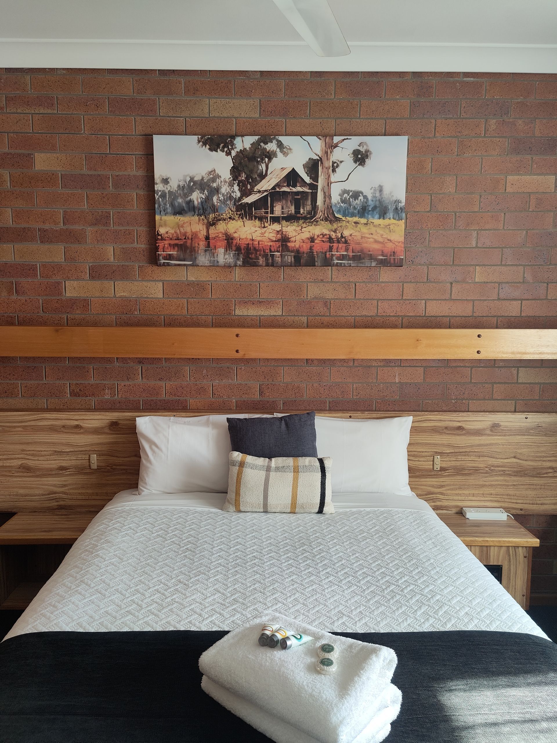 Cozy bedroom with brick wall, landscape art, double bed, white bedding, and a towel laid across the foot of the bed