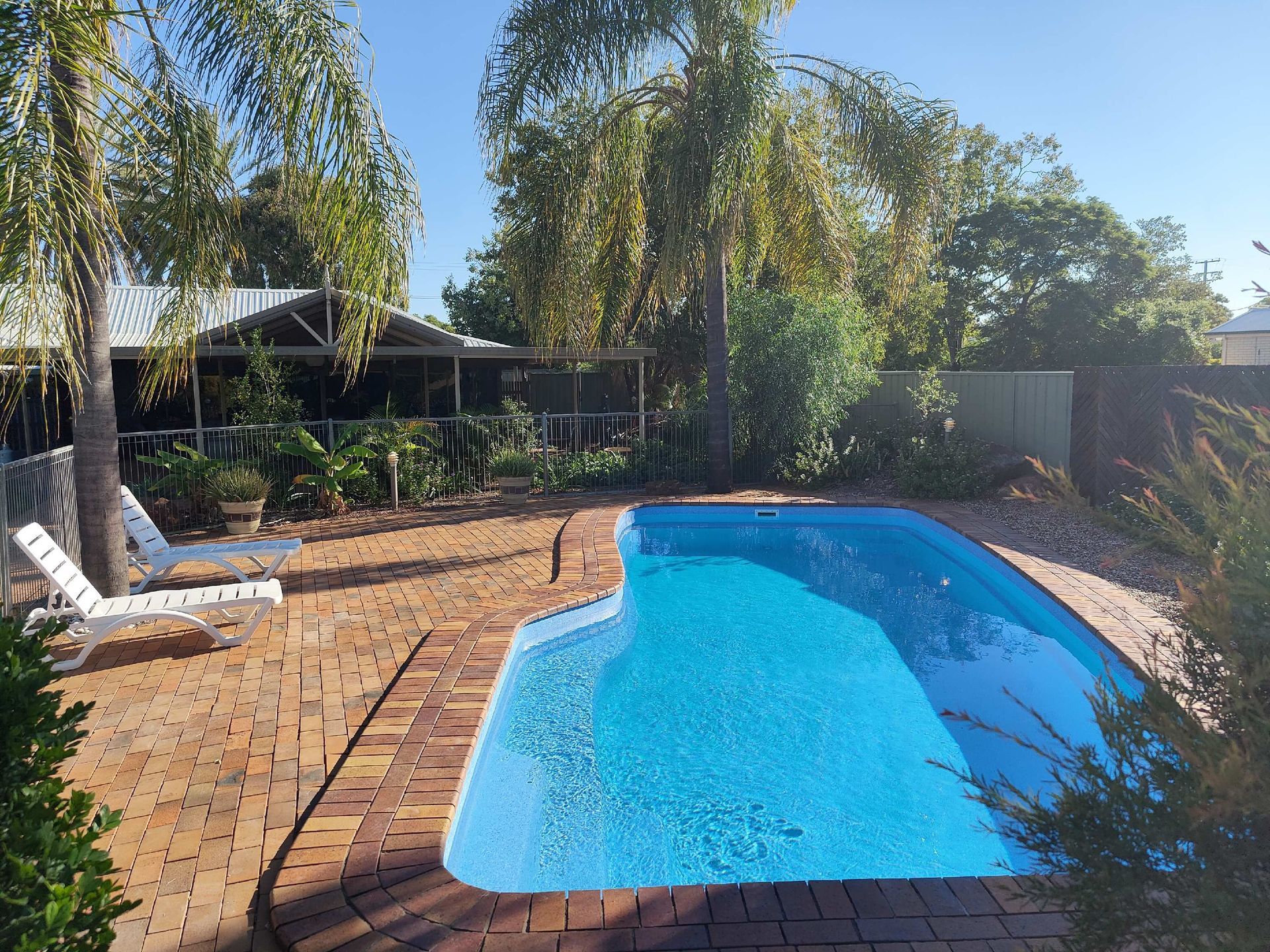 Backyard pool with blue water, brick patio, palm trees, and lounge chair in sunny weather