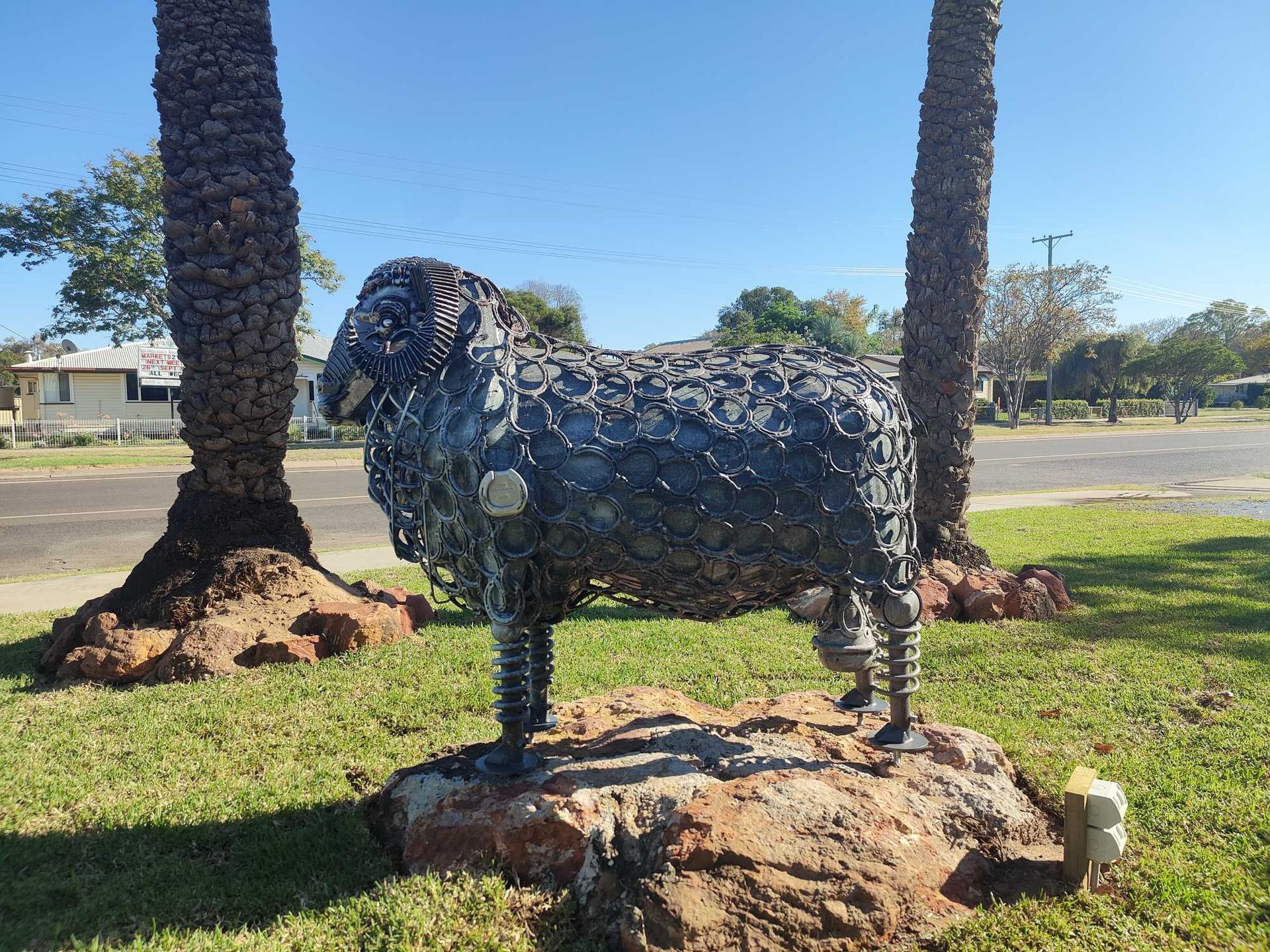 Metal animal sculpture on a grassy roadside with palm trees in the background.