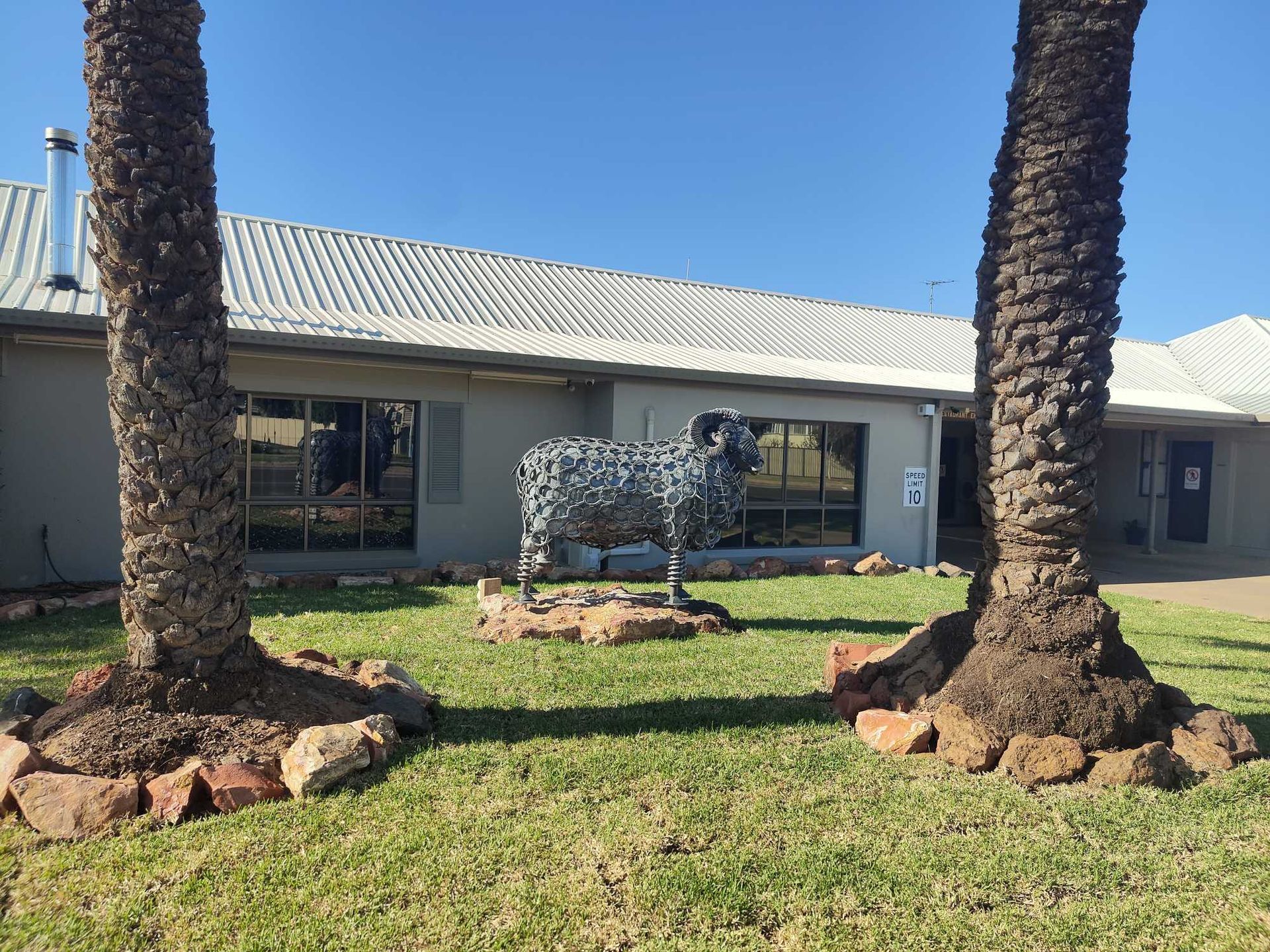 Single-story house with white roof, green lawn, and two palm trees framing a decorative cow sculpture.