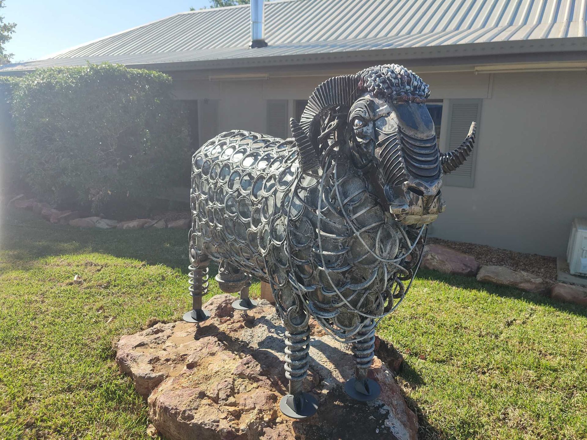 Metal sheep sculpture in a grassy yard beside a beige building, standing on a rock base.