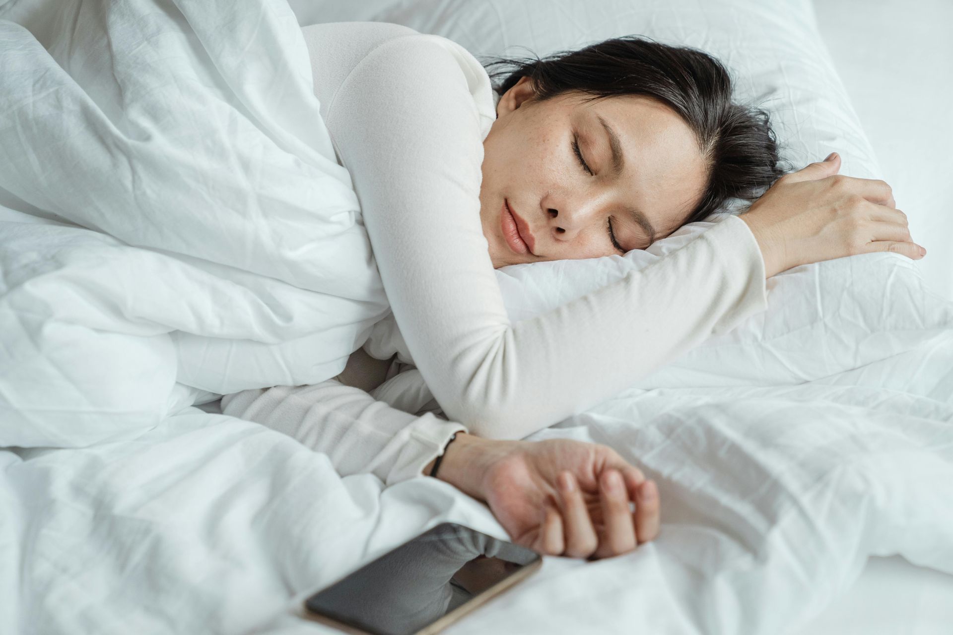Person sleeping in white bedding with a smartphone on the pillow beside them.