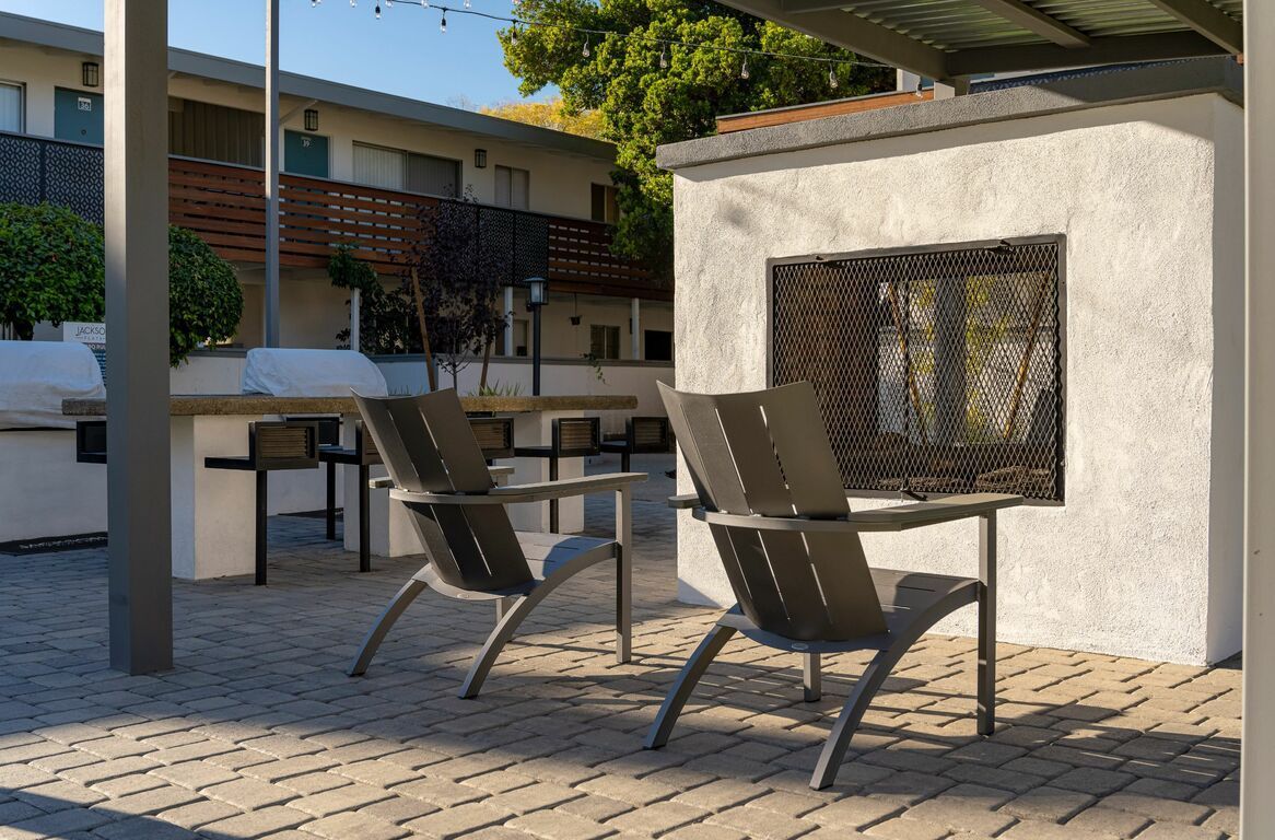 Two lounge chairs on a paved patio beside a white fireplace under a shaded pergola.