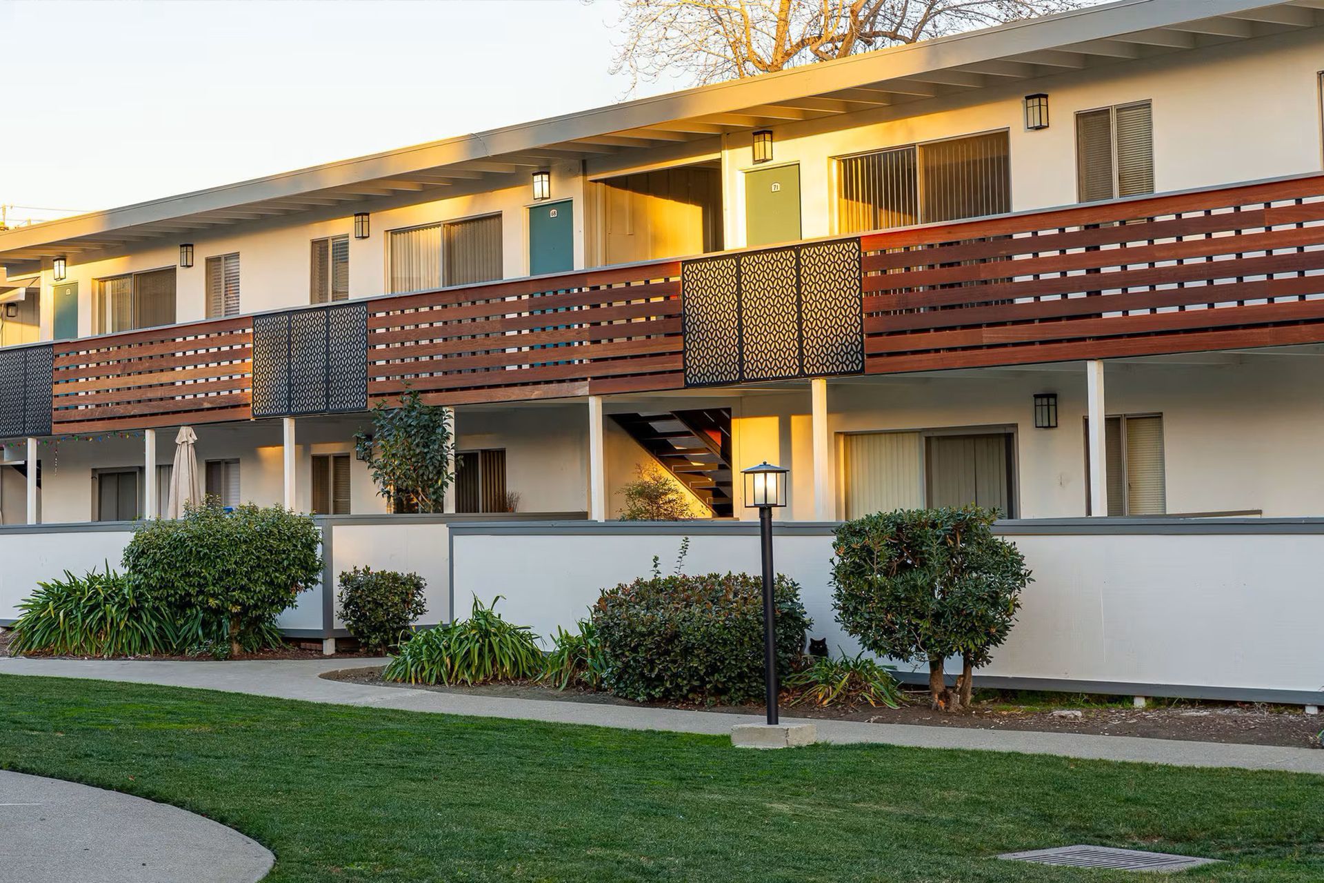 Two-story apartment building with brown balcony railings and white walls, seen across a grassy lawn.