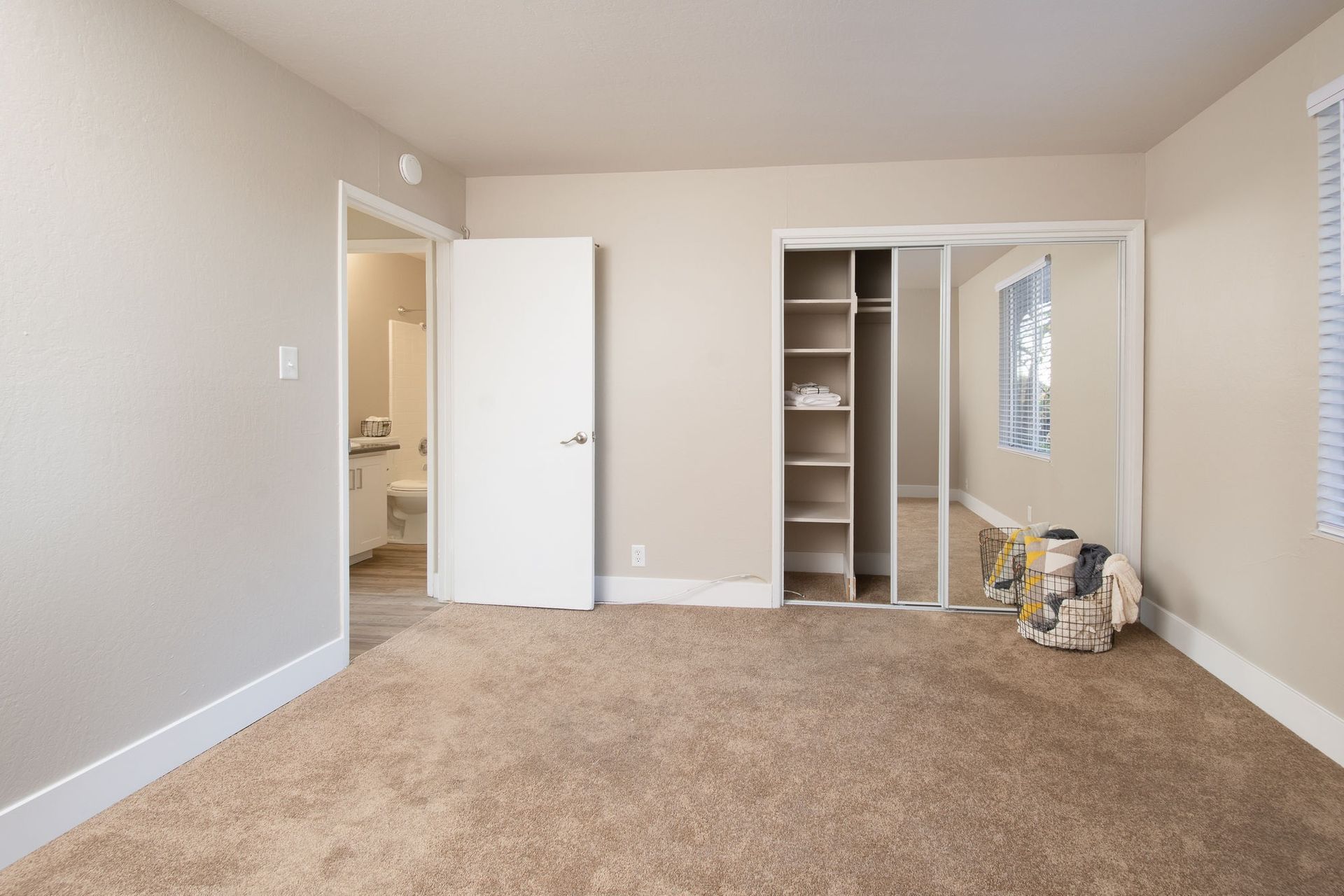 Empty beige bedroom with carpet, open closet doors, and a doorway to a bathroom.