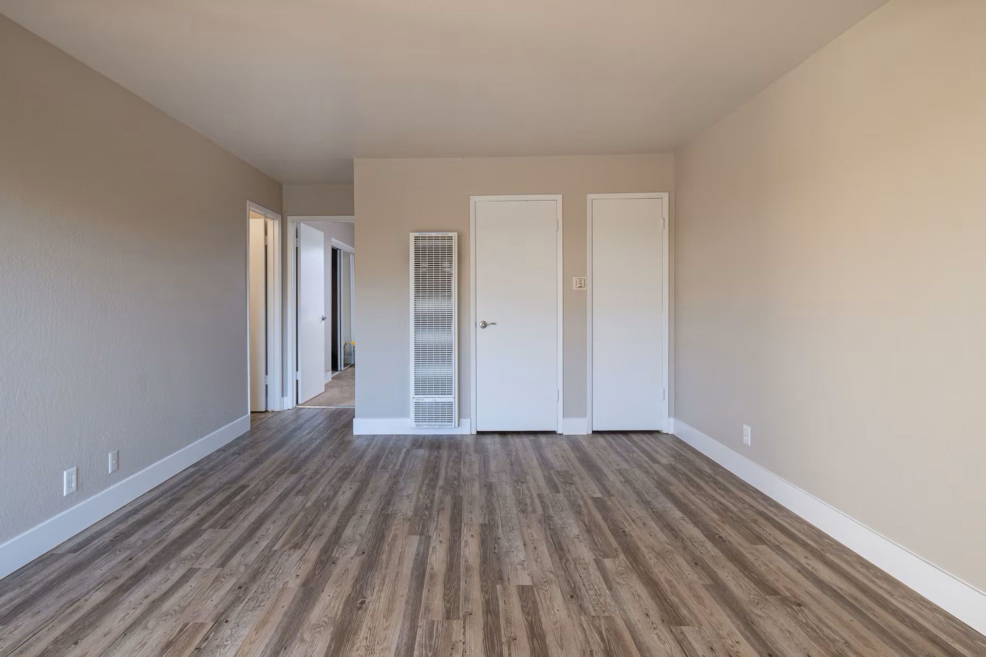 Empty beige living room with wood-look floors, white doors, and a narrow hallway view
