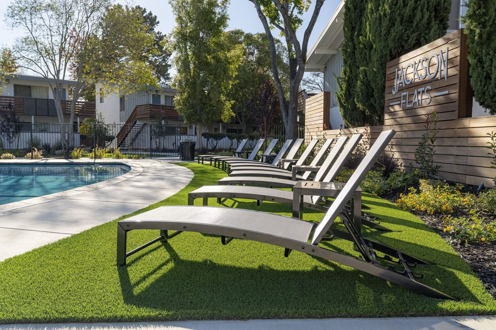 Poolside lounge chairs on bright green turf beside a swimming pool, with trees and a wooden wall.