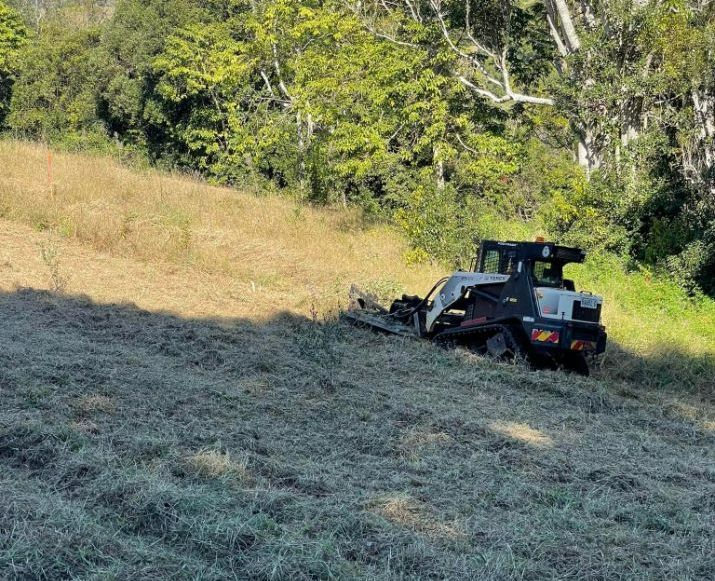 A Bulldozer is Cutting Grass in a Field With Trees in the Background — Tallow Tree Services Pty Ltd in Kyogle, NSW