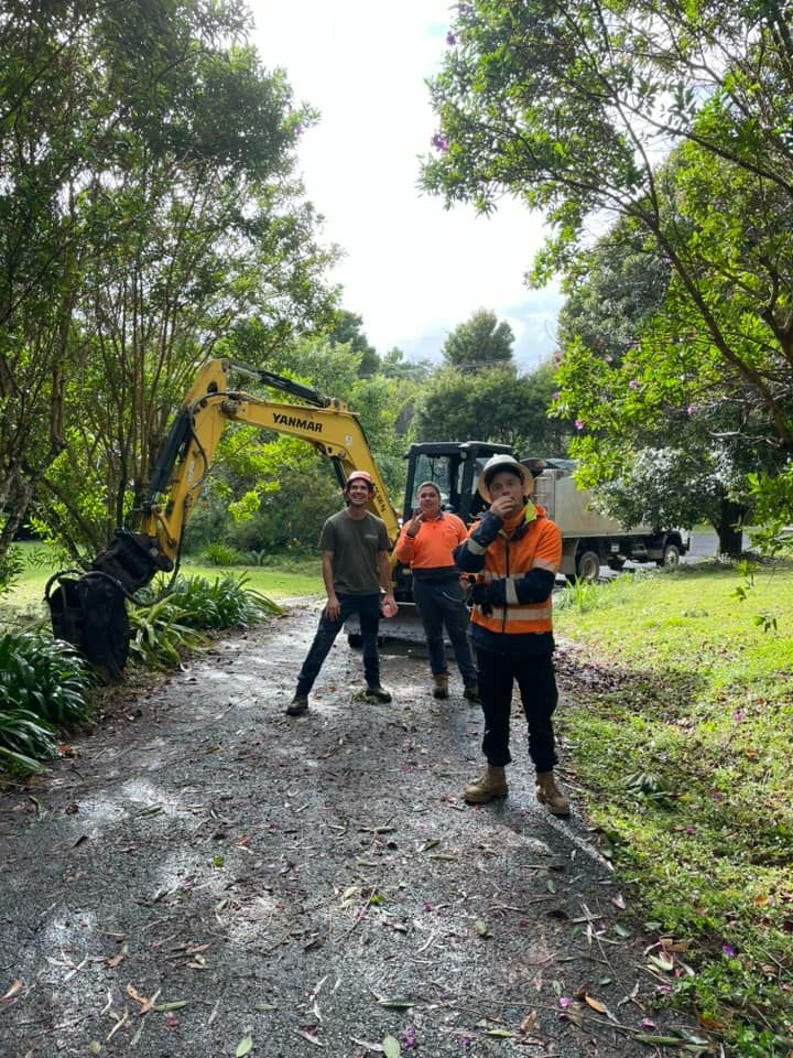 A Group of Men Are Standing Next to a Yellow Excavator on a Dirt Road — Tallow Tree Services Pty Ltd in Mullumbimby, NSW