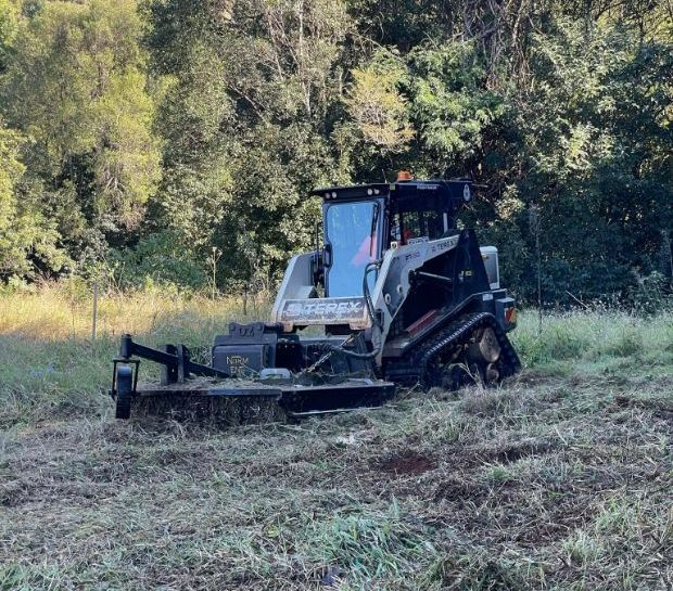 A Tractor is Cutting Grass in a Field With Trees in the Background — Tallow Tree Services Pty Ltd in Mullumbimby, NSW