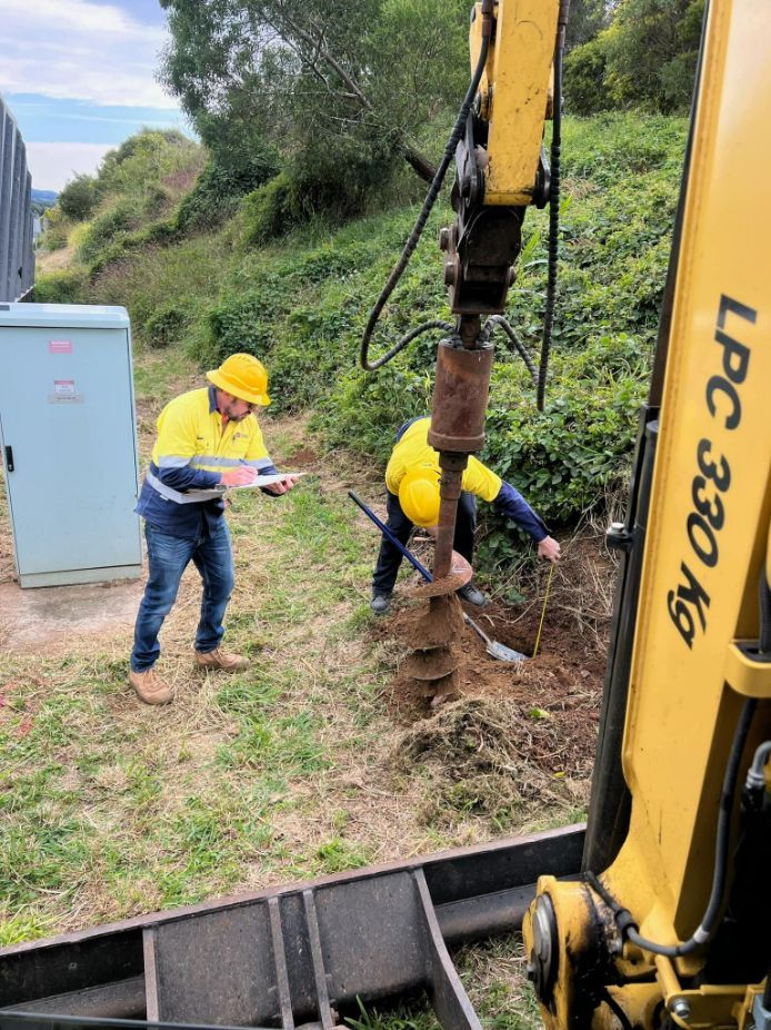 Two Men Are Digging a Hole in the Ground Next to a Yellow Excavator — Tallow Tree Services Pty Ltd in Bangalow, NSW