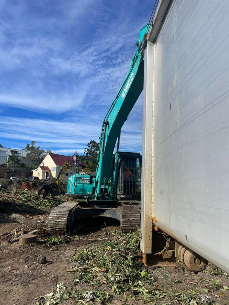 A Green Excavator is Standing Next to a White Building — Tallow Tree Services Pty Ltd in Yamba, NSW