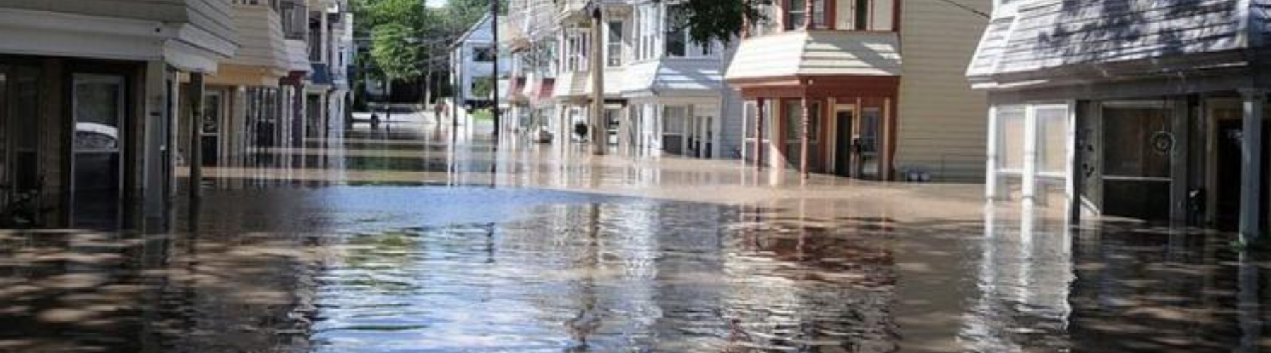 A flooded street with buildings and trees reflected in the water.