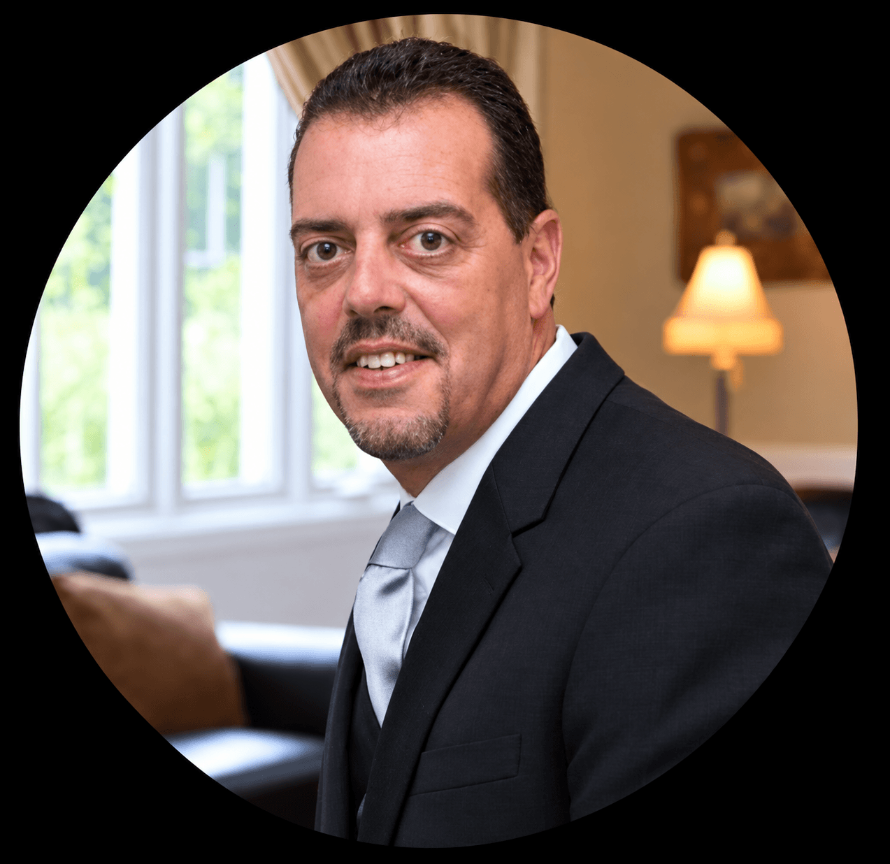 Jim Chanske of Insurance Warehouse in a black suit and silver tie smiling in a warmly lit room near a window.