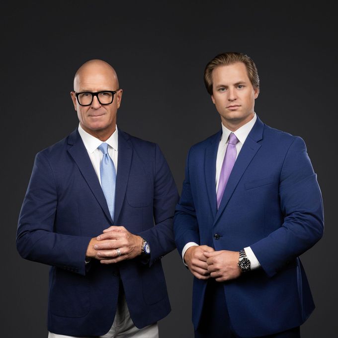 Two men in blue suits posing against a dark background, one bald with glasses and one with styled hair.