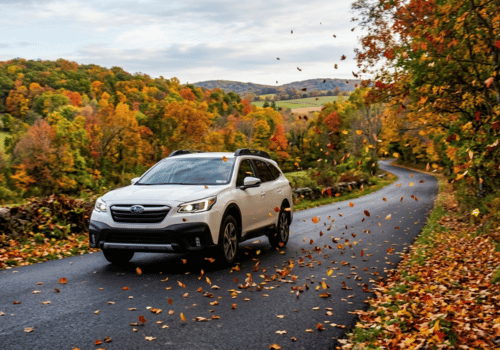 White Subaru Outback driving along a winding road surrounded by colorful fall foliage