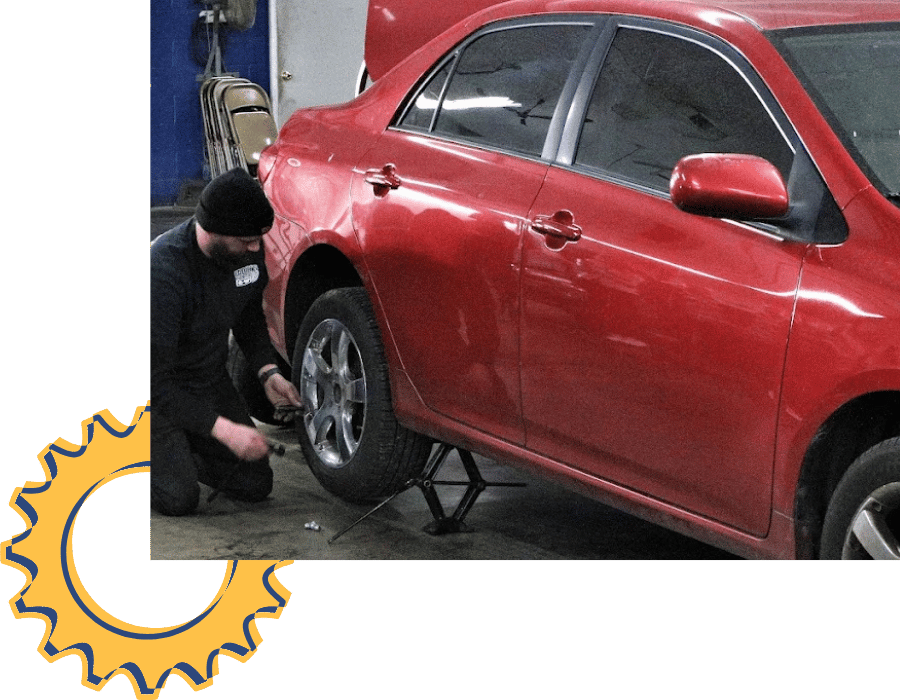 Mechanic changing tire on red sedan inside auto repair shop.