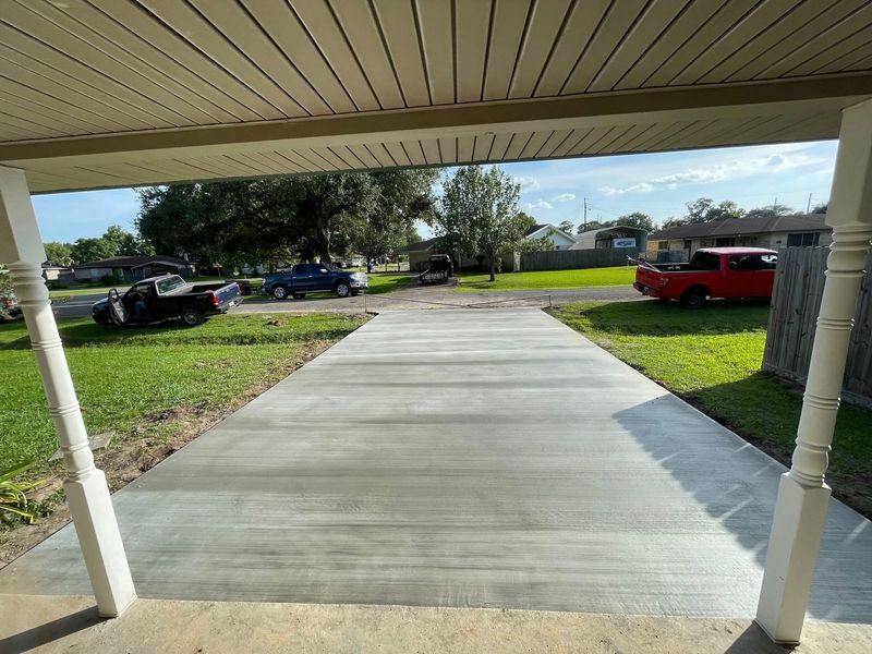 View from a porch of a newly poured concrete driveway, with cars and grass in the distance.