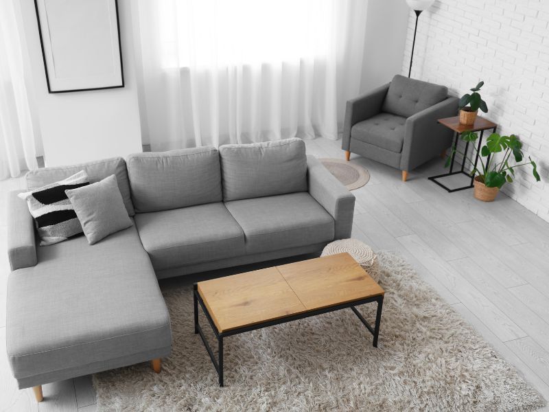 Bright living room with gray sectional sofa, wooden coffee table, armchair, plants, and sheer curtains