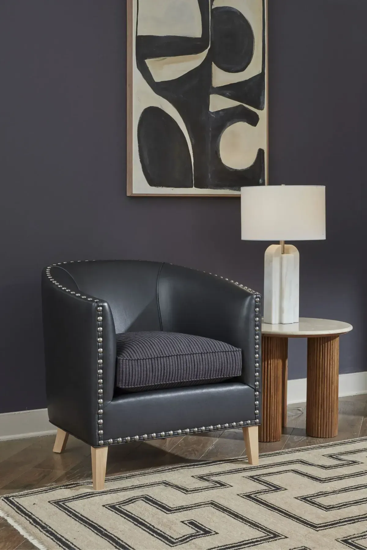 Modern living room corner with black armchair, round side table, lamp, abstract wall art, and patterned rug.