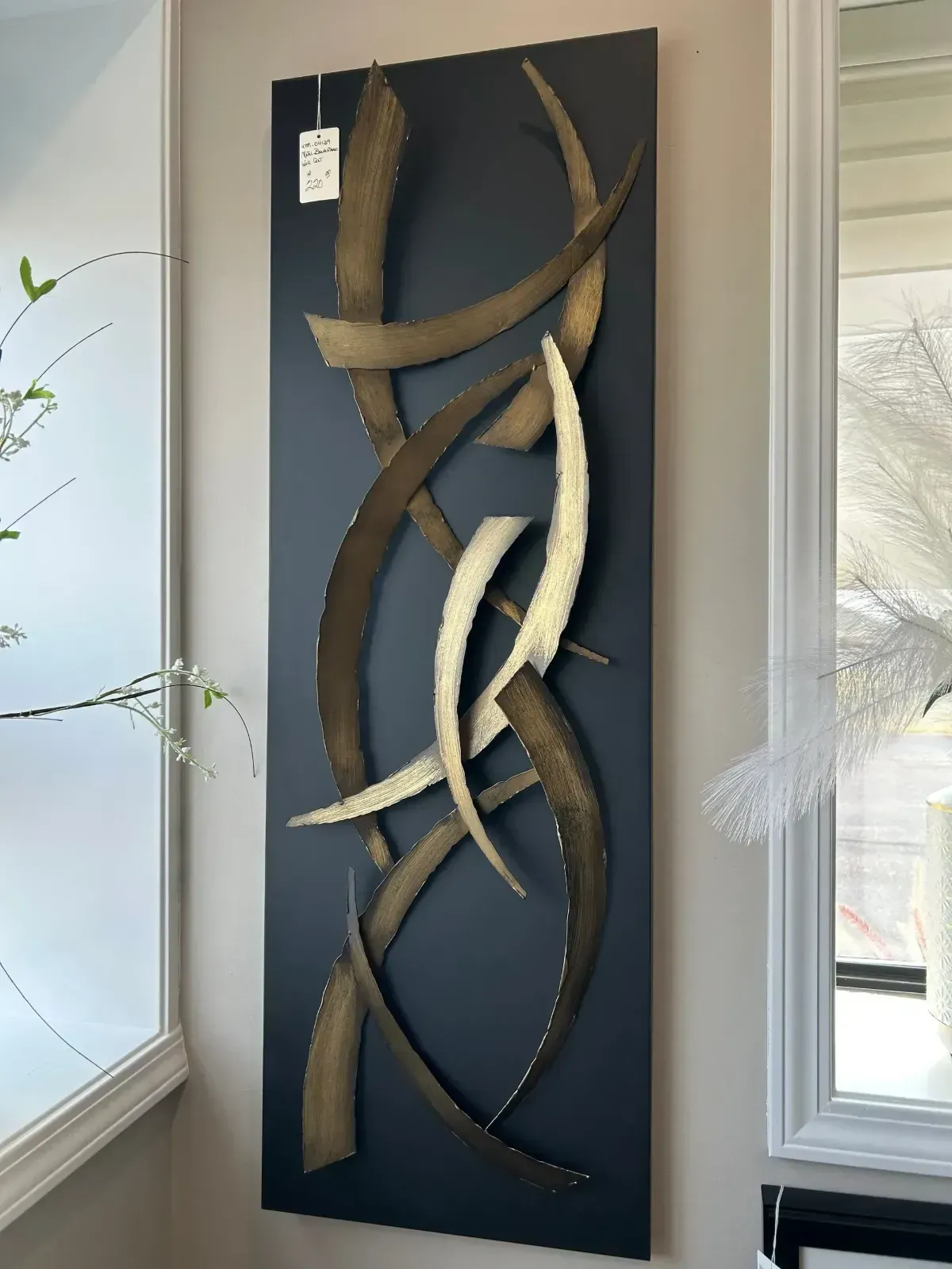 Tall abstract metal wall sculpture with curved bronze and cream ribbons on a dark panel by a window