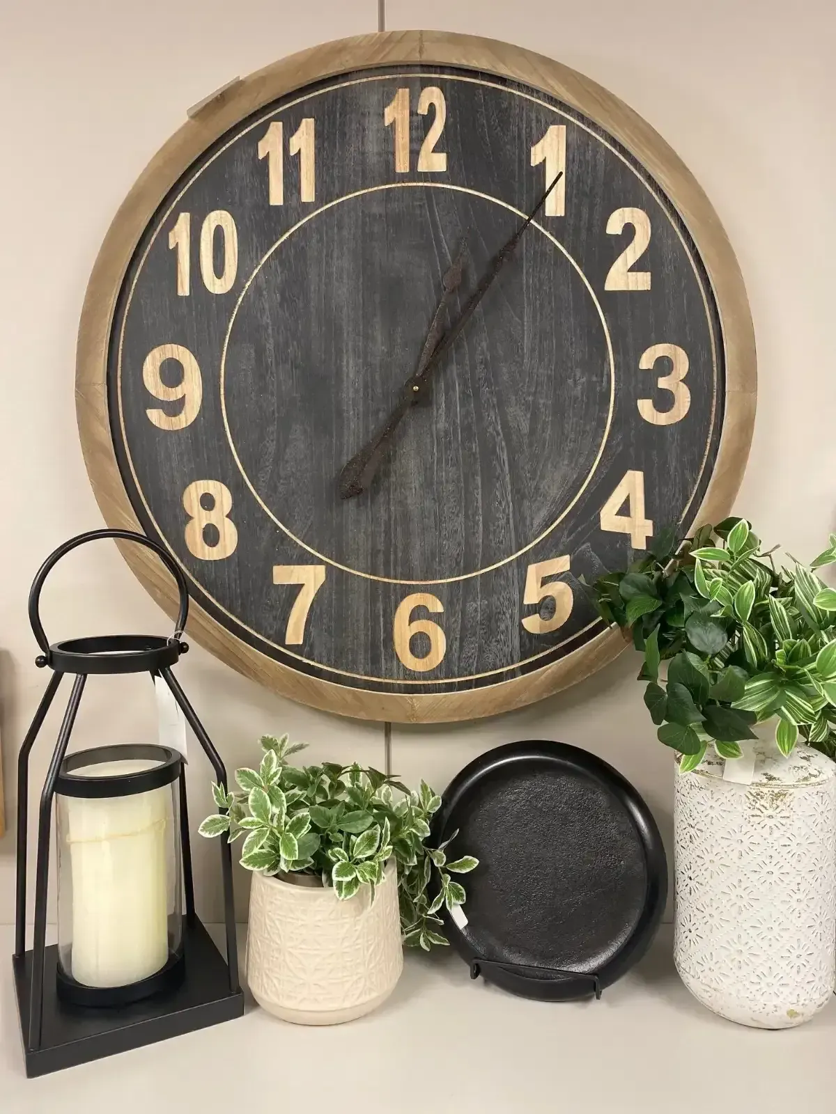 Rustic wall clock above candles, potted plants, and a black plate on a shelf arrangement